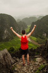 Young man in red sports shirt raises his arms victoriously after reaching the top of a viewpoint in Tam Coc