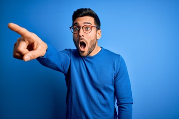 Young handsome man with beard wearing casual sweater and glasses over blue background Pointing with...