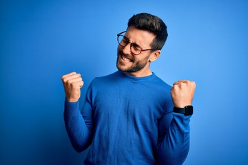 Young handsome man with beard wearing casual sweater and glasses over blue background very happy...