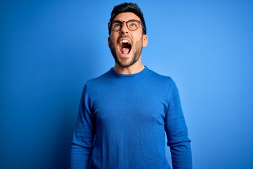 Young handsome man with beard wearing casual sweater and glasses over blue background angry and mad screaming frustrated and furious, shouting with anger. Rage and aggressive concept.