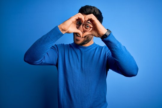 Young handsome man with beard wearing casual sweater and glasses over blue background Doing heart shape with hand and fingers smiling looking through sign