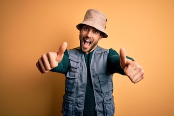 Handsome tourist man with beard on vacation wearing explorer hat over yellow background approving doing positive gesture with hand, thumbs up smiling and happy for success. Winner gesture.