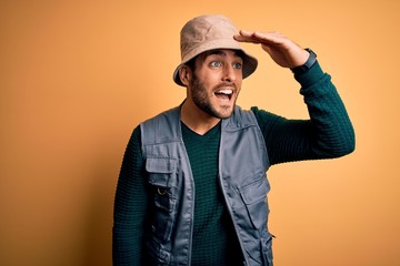 Handsome tourist man with beard on vacation wearing explorer hat over yellow background very happy and smiling looking far away with hand over head. Searching concept.