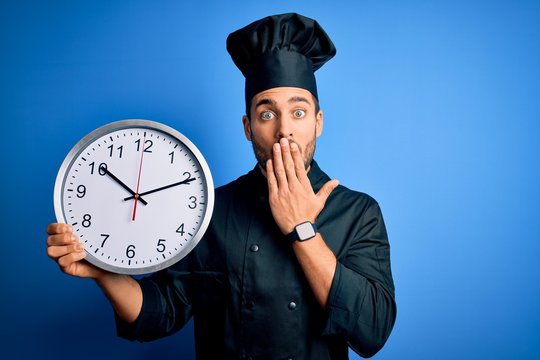 Young handsome cooker man with beard wearing uniform holding clock doing countdown cover mouth with hand shocked with shame for mistake, expression of fear, scared in silence, secret concept
