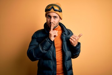 Young handsome skier man with beard wearing snow sportswear and ski goggles asking to be quiet with finger on lips pointing with hand to the side. Silence and secret concept.