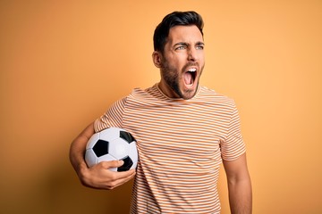 Handsome player man with beard playing soccer holding footballl ball over yellow background angry...