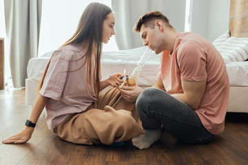 caucasian couple sit on the floor at home, woman help to light a bong bowl with cannabis. they use...