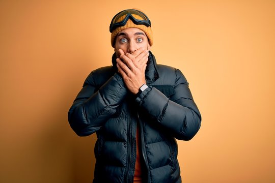 Young handsome skier man with beard wearing snow sportswear and ski goggles shocked covering mouth with hands for mistake. Secret concept.