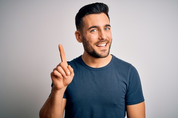 Young handsome man wearing casual t-shirt standing over isolated white background showing and pointing up with finger number one while smiling confident and happy.