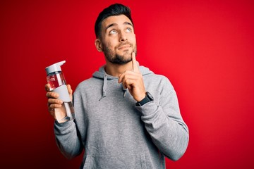 Young handsome sportsman drinking bottle of water to refeshment over red background serious face...