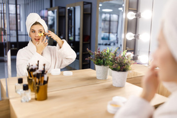 Young beautiful girl in a white robe and with a towel on her head puts patches under her eyes in front of a mirror. Skin care.