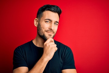 Young handsome man wearing casual black t-shirt standing over isolated red background looking confident at the camera with smile with crossed arms and hand raised on chin. Thinking positive.