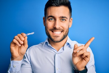 Young handsome man holding dental aligner tooth correction over blue background very happy pointing with hand and finger to the side