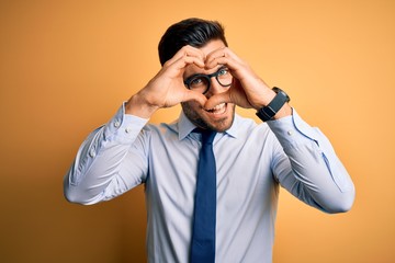 Young handsome businessman wearing tie and glasses standing over yellow background Doing heart shape with hand and fingers smiling looking through sign