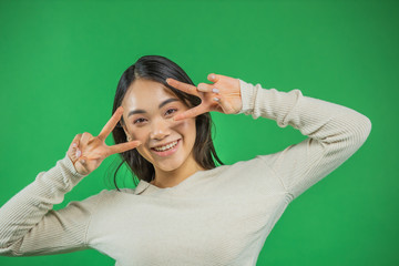 Elegant graceful glamor, attractive pretty calm Asian lady, isolated on a background of green glitter holding her v sign on two hands near her eyes, makes a lovely radiant smile