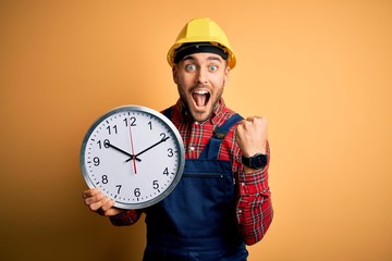 Young builder man wearing safety helmet holding big clock over yellow background screaming proud...