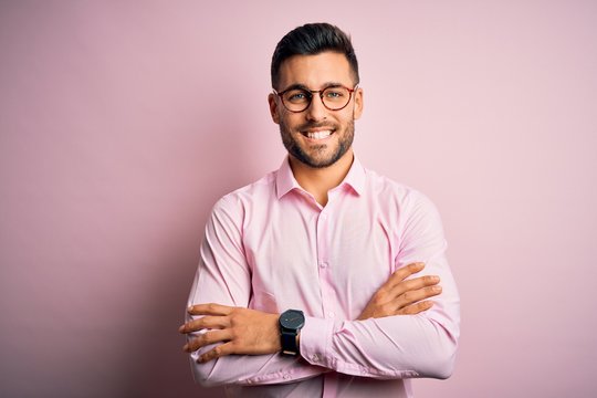 Young handsome man wearing elegant shirt and glasses standing over pink background happy face smiling with crossed arms looking at the camera. Positive person.