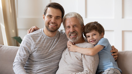 Portrait of three generations of men sit rest on couch in living room hugging, happy little boy with young dad and senior grandfather relax on sofa at home, enjoy family leisure weekend together
