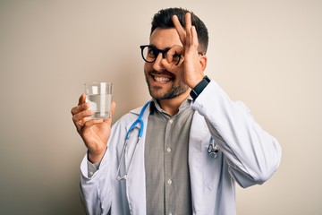 Young doctor man wearing stethoscope drinking a fresh glass of water over isolated background with happy face smiling doing ok sign with hand on eye looking through fingers