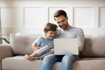 Smiling little boy relax on couch at home with young dad watch funny videos on laptop together,...