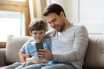 Happy young father sit relax on couch in living room with little son make selfie on smartphone, dad...