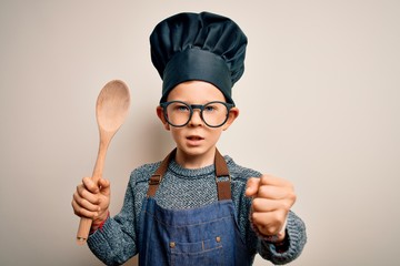 Young little caucasian cook kid wearing chef uniform and hat cooking using wooden spoon annoyed and...