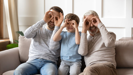 Naklejka na ściany i meble Portrait of overjoyed three generations of men sit on couch posing for funny picture together, smiling little boy with young dad and senior grandfather make face gestures look at camera at home