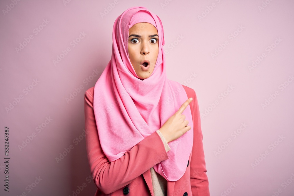 Poster young beautiful girl wearing muslim hijab standing over isolated pink background surprised pointing 