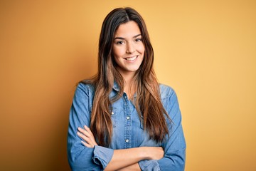 Young beautiful girl wearing casual denim shirt standing over isolated yellow background happy face smiling with crossed arms looking at the camera. Positive person.