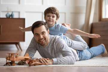 Portrait of cute little boy lying with smiling young father on floor engaged in funny game together, happy small preschooler son playing with loving dad, enjoying family leisure weekend at home