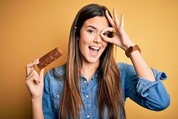 Young beautiful girl holding healthy protein bar standing over isolated yellow background with...