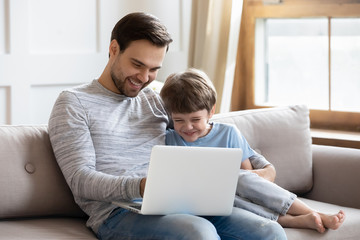 Overjoyed young dad sit relax on couch with happy little son have fun using laptop together, smiling father rest on sofa at home with small preschooler boy child laugh watch funny video on computer