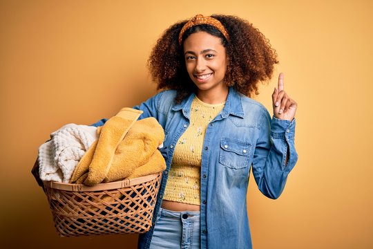 Young african american woman with afro hair holding wicker basket with clothes surprised with an idea or question pointing finger with happy face, number one