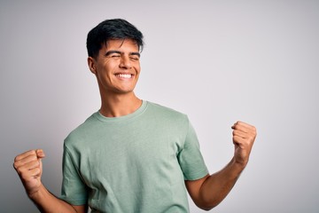 Young handsome man wearing casual t-shirt standing over isolated white background very happy and excited doing winner gesture with arms raised, smiling and screaming for success. Celebration concept.