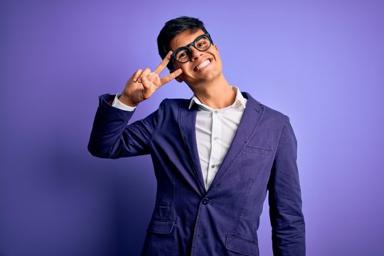 Young handsome business man wearing jacket and glasses over isolated purple background Doing peace symbol with fingers over face, smiling cheerful showing victory