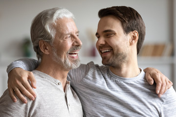 Overjoyed young millennial man embrace mature dad have fun talk and laugh at home, happy elderly...