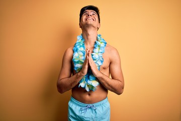 Young handsome tourist man on vacation wearing swimwear and hawaiian lei flowers begging and praying with hands together with hope expression on face very emotional and worried. Begging.