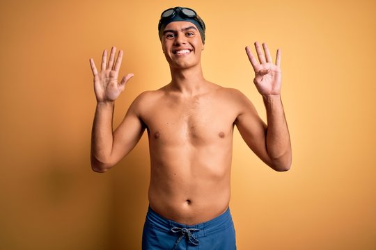 Young handsome man shirtless wearing swimsuit and swim cap over isolated yellow background showing and pointing up with fingers number nine while smiling confident and happy.