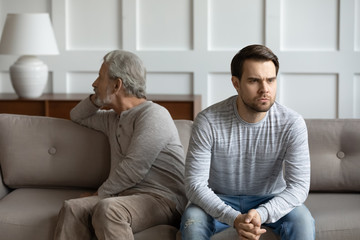Upset stubborn young man and elderly father sit separately on couch at home avoid talking after fight, offended mad mature dad have misunderstanding with adult grown-up son, generation gap concept