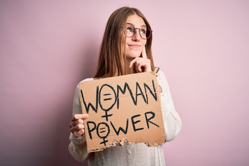 Young beautiful redhead woman asking for women rights holding banner over pink background serious...