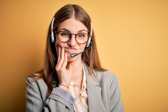 Young redhead call center agent woman overworked wearing glasses using headset thinking looking tired and bored with depression problems with crossed arms.