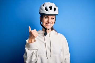 Young beautiful redhead cyclist woman wearing bike helmet over isolated blue background doing happy thumbs up gesture with hand. Approving expression looking at the camera showing success.