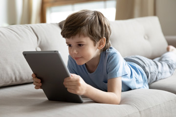 Smart small preschooler boy lying on couch in living room watch video cartoon on tablet, little child relax on sofa at home using modern pad device learning, kids and technology, education concept