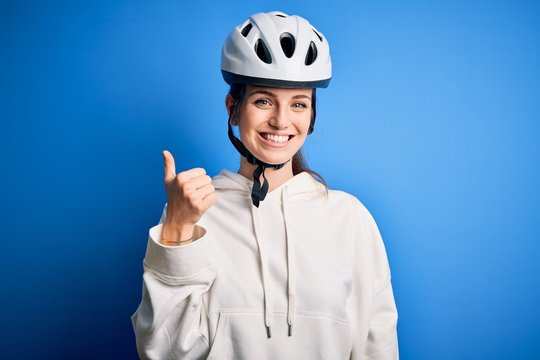 Young beautiful redhead cyclist woman wearing bike helmet over isolated blue background doing happy thumbs up gesture with hand. Approving expression looking at the camera showing success.