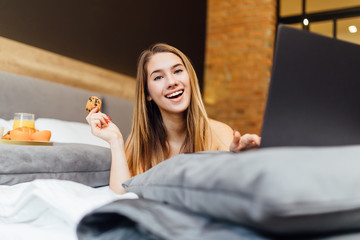 Photo of a happy blonde woman have use laptop on bedroom with cake on hand.