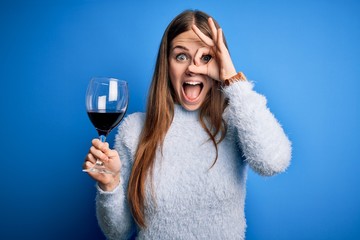 Young beautiful redhead woman drinking glass of red wine over isolated blue background with happy...