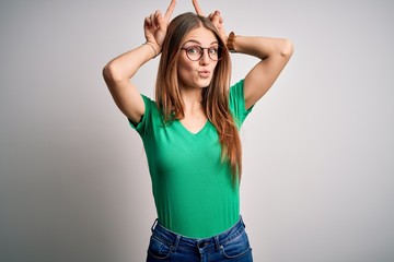 Obraz na płótnie Canvas Young beautiful redhead woman wearing casual green t-shirt and glasses over white background doing funny gesture with finger over head as bull horns