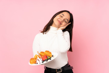 Young brunette woman holding waffles over isolated pink background making sleep gesture in dorable expression