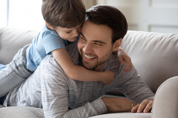 Cute little boy lying on happy young father hugging relaxing together on sofa in living room, overjoyed small preschooler son embrace loving dad, show affection, enjoy family weekend at home