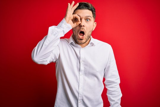 Young business man with blue eyes wearing elegant shirt standing over red isolated background doing ok gesture shocked with surprised face, eye looking through fingers. Unbelieving expression.